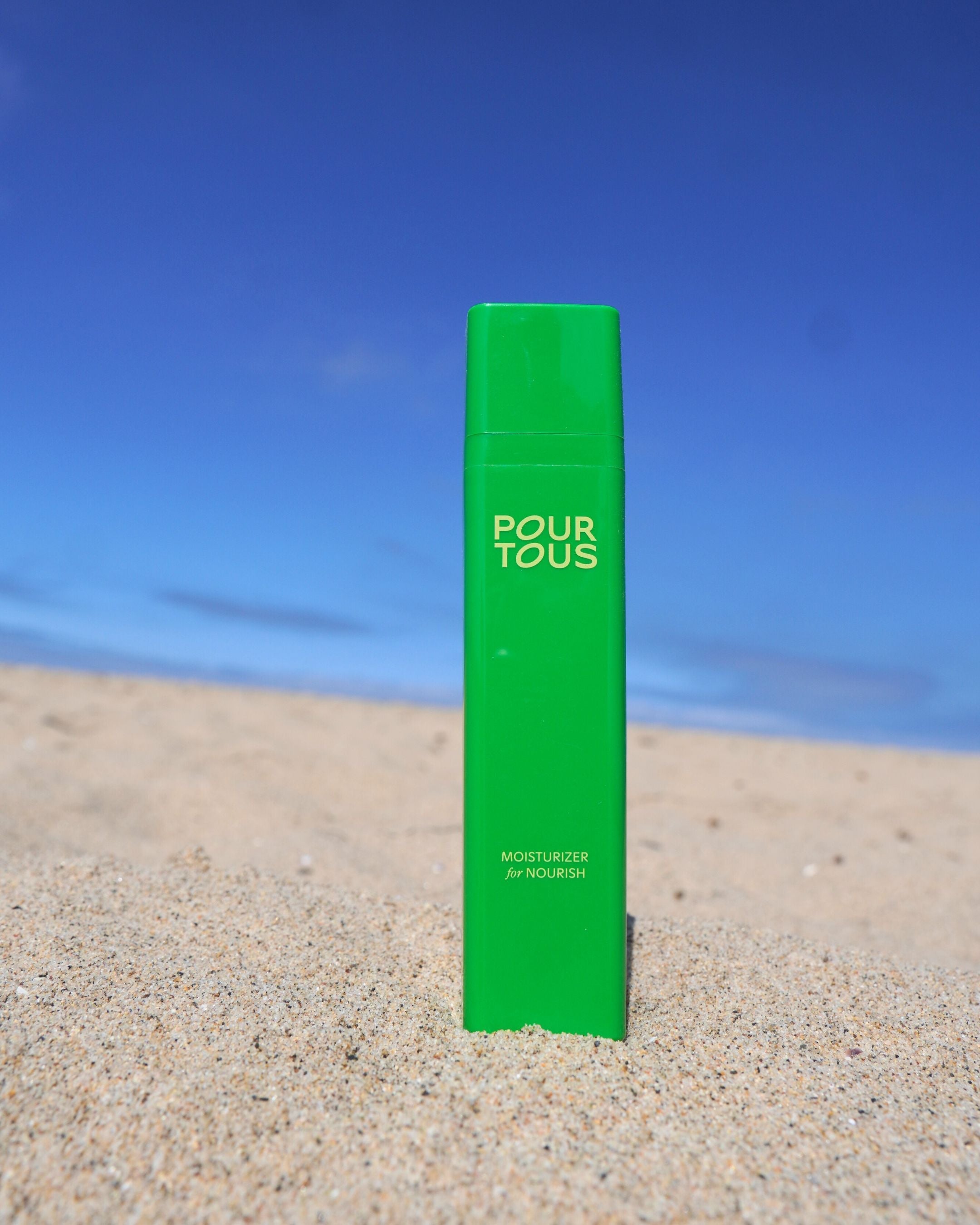 Green moisturizer bottle in sand under blue sky.