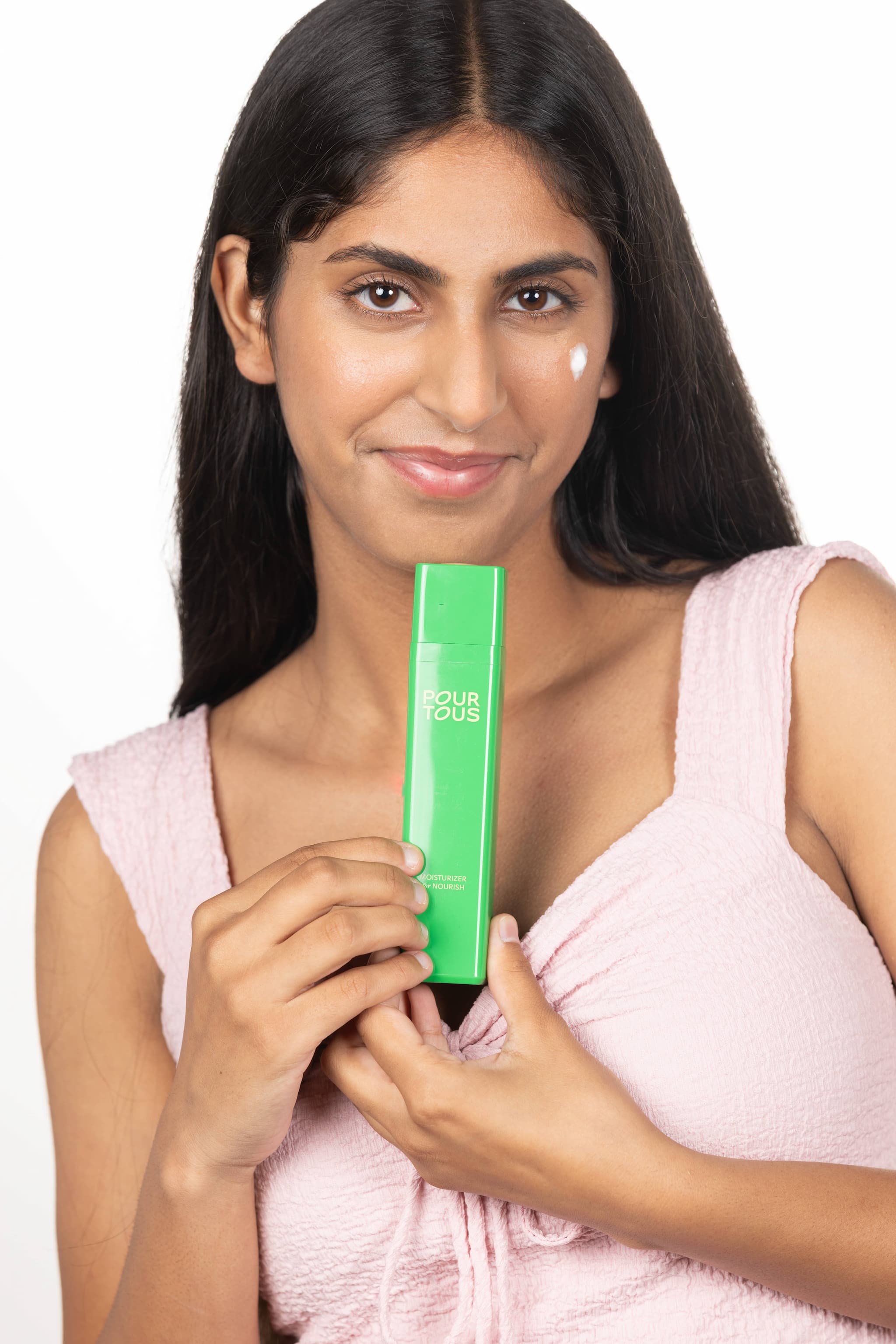 Person holding green skincare product labeled "Pour Tous" with cream on cheek.