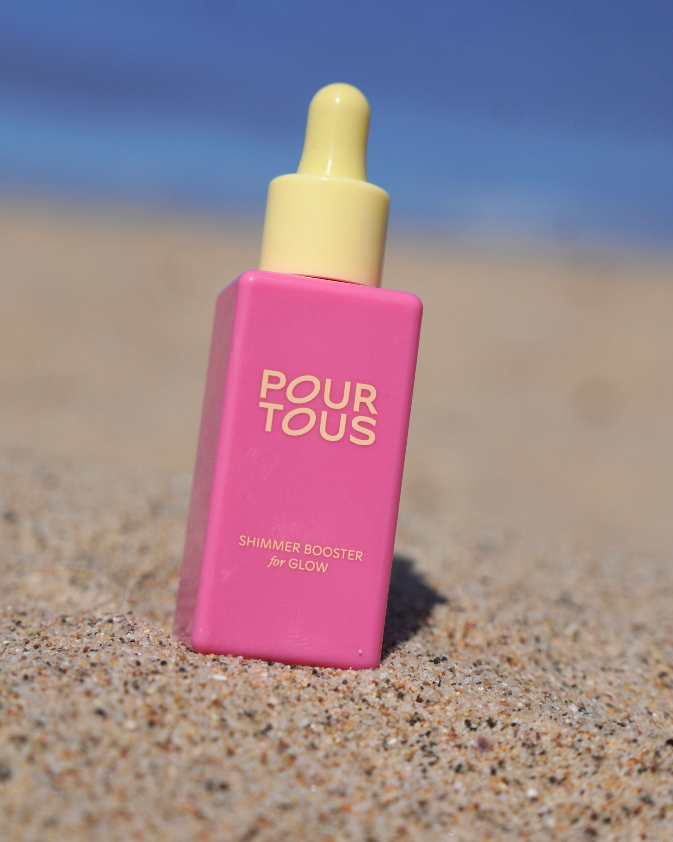 Pink shimmer booster bottle on sand with blue sky background.