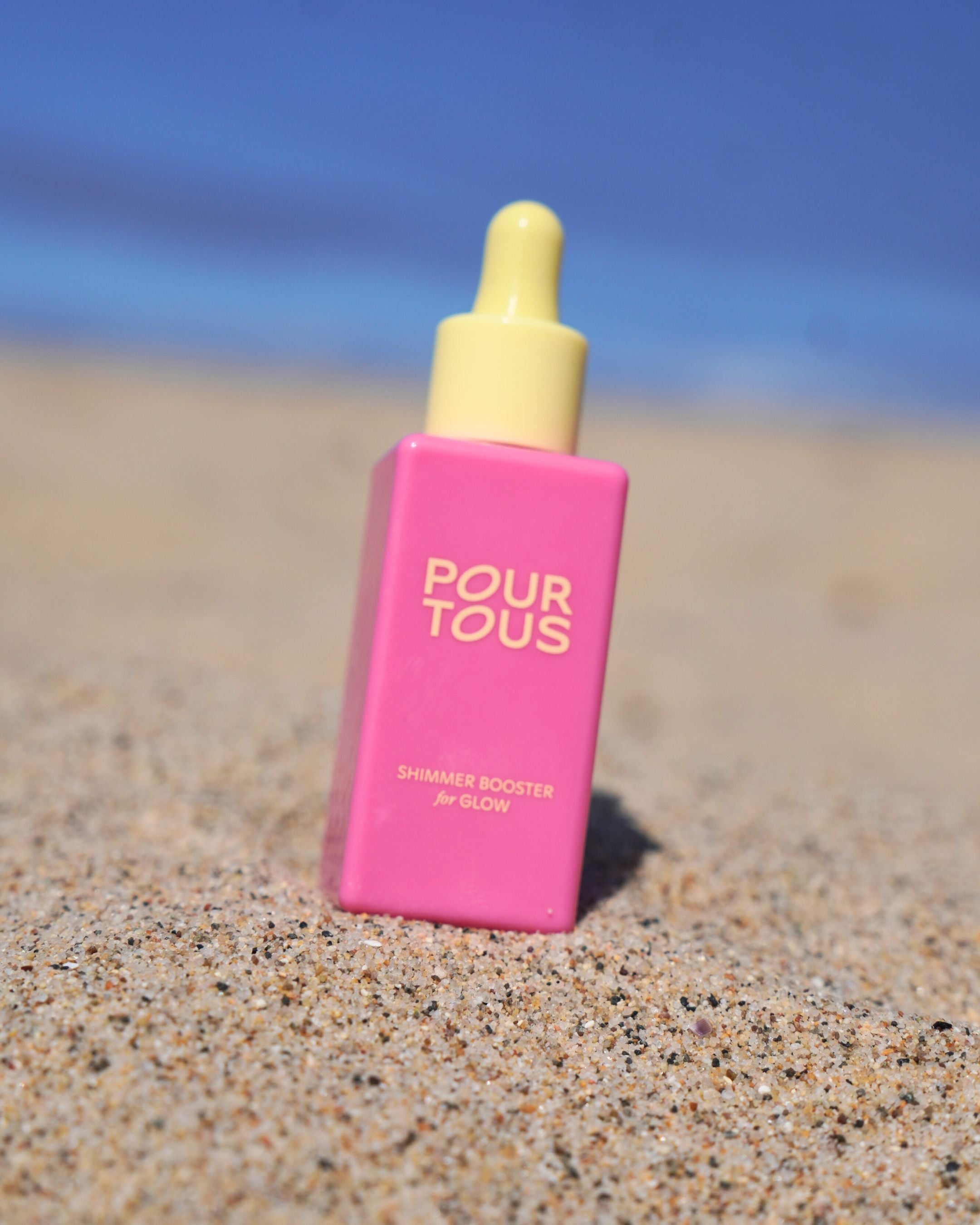 Pink shimmer booster bottle on sandy beach with blue sky background.